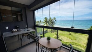 A balcony with a view of the ocean, a small table with chairs, and a kitchen area with a coffee maker. - Kauai Praia dos Carneiros |Apartamento Pé na Areia (Tamandaré)