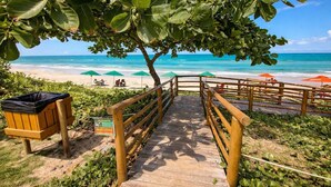 A wooden pathway leading to a beach with umbrellas and a trash bin. - Kauai Praia dos Carneiros |Apartamento Pé na Areia (Tamandaré)