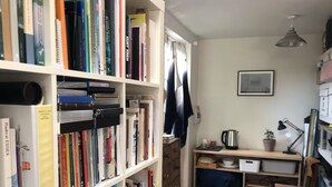 Un couloir étroit avec une bibliothèque remplie de livres, un petit coin cuisine avec une bouilloire, un bureau et une chaise.