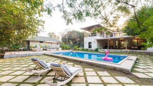 A swimming pool with lounge chairs and a flamingo float, surrounded by a garden and a house. - ELIVAAS Casa Mocha Exquisite 5BHK Villa (Alibag)