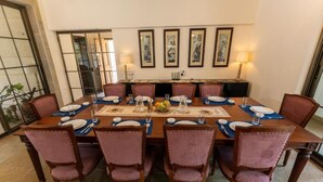 A dining room with a large table set for eight, surrounded by six chairs, with artwork on the walls and a view through a glass door. - ELIVAAS Casa Mocha Exquisite 5BHK Villa (Alibag)