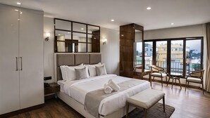 A modern hotel bedroom with a large bed, a desk, and a balcony with chairs. - Valmere Chery Blossom Suites (Shillong)