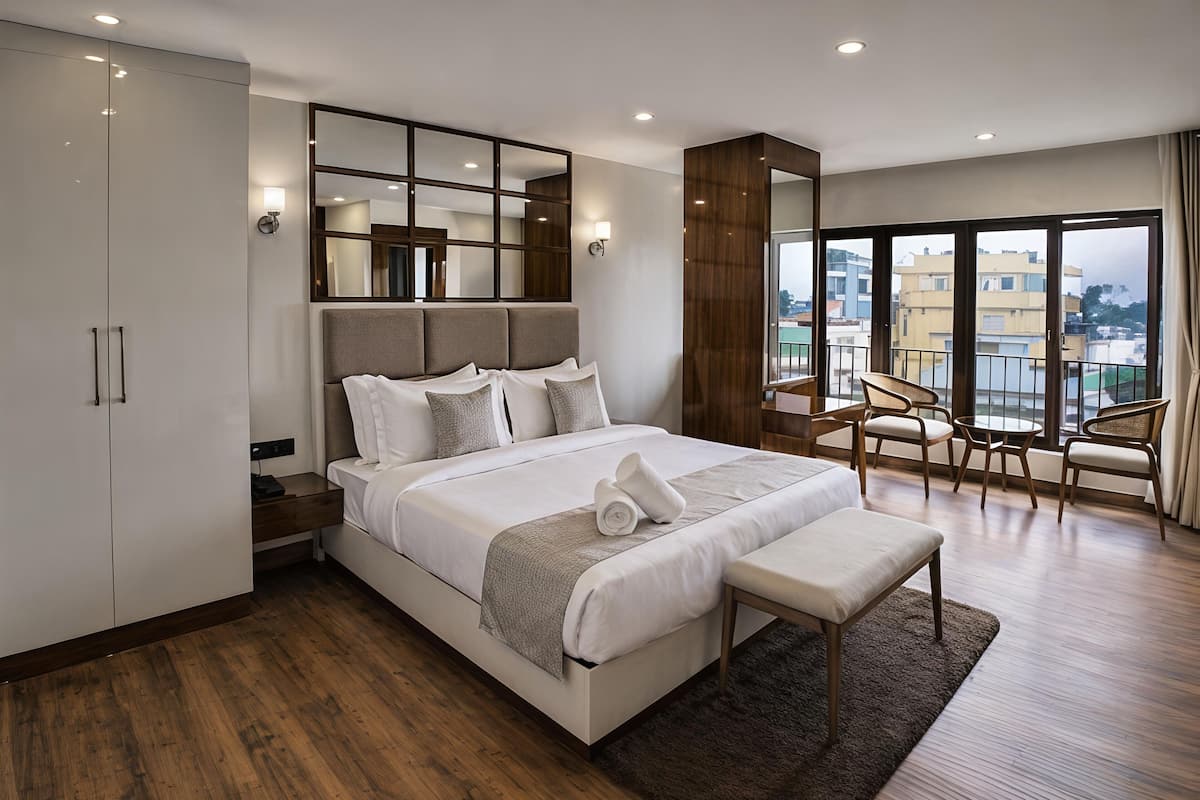 A modern hotel bedroom with a large bed, a desk, and a balcony with chairs.