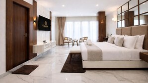 A modern hotel room with a large bed, a TV mounted on the wall, a seating area with chairs, and a large window with curtains. - Valmere Chery Blossom Suites (Shillong)