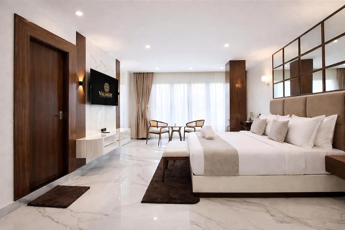 A modern hotel room with a large bed, a TV mounted on the wall, a seating area with chairs, and a large window with curtains.
