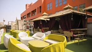 Outdoor wicker furniture on artificial grass, a building with red walls, and a traditional structure with a thatched roof. - First Impression (Varanasi)