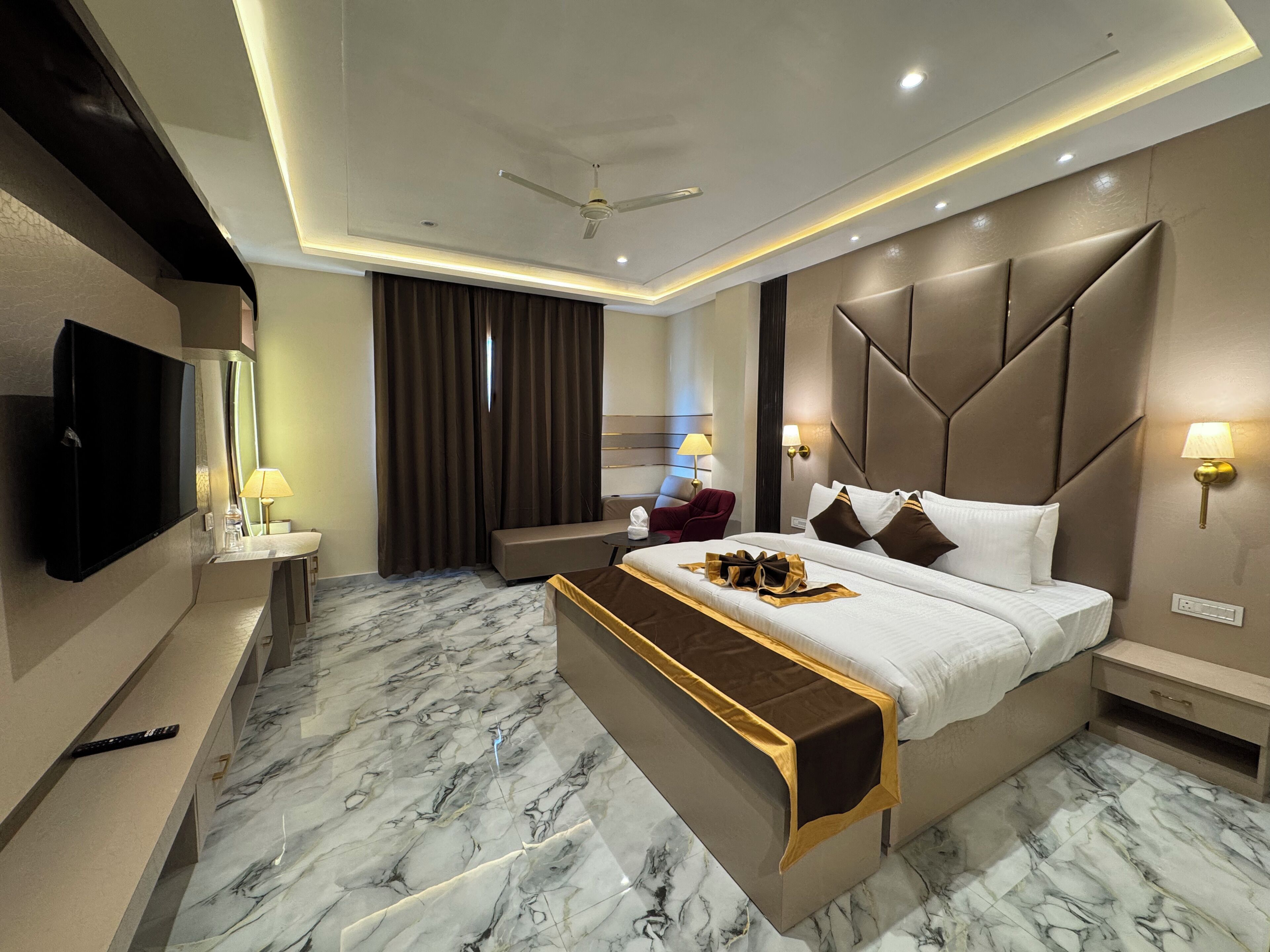 A modern hotel room with a large bed, a flat-screen TV, a sofa, a desk, and a ceiling fan.