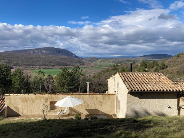 Un bâtiment rustique avec un toit en tuiles, un parasol blanc et une vue sur des collines ondoyantes et une montagne au loin.