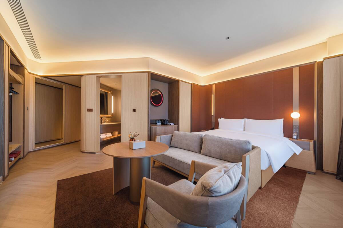 A hotel room with a bed, a sofa, a small table, a chair, a round mirror, and a wall-mounted light fixture.