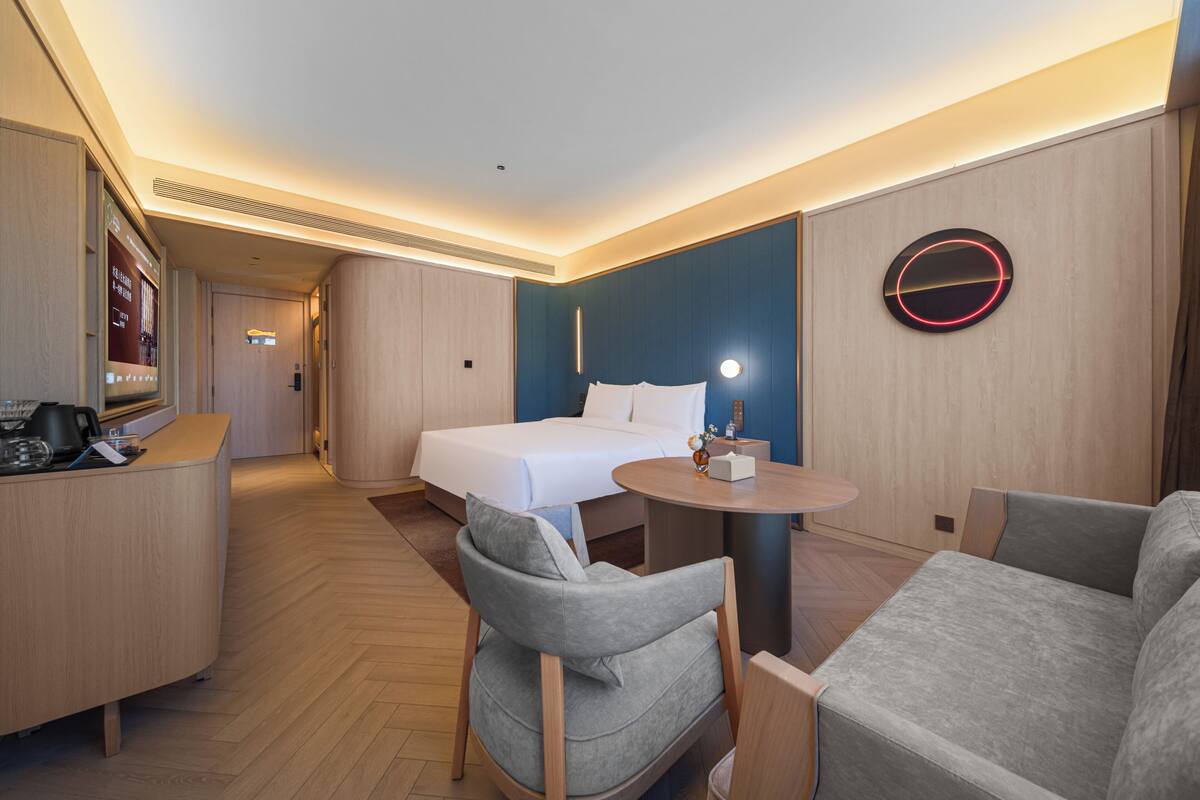 A hotel room with a bed, a desk, a chair, a round table, and a wall-mounted light fixture.