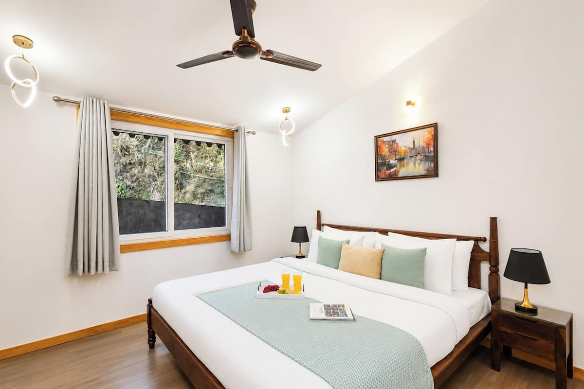 A hotel room with a bed, a ceiling fan, a window with curtains, a bedside table with a lamp, and a painting on the wall.
