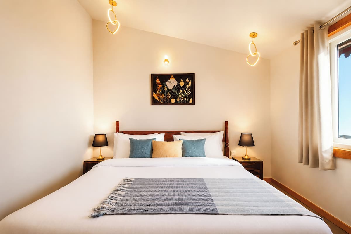 A neatly made bed with a patterned throw, two bedside tables with lamps, a wall-mounted artwork, and a window with curtains.