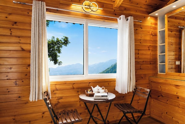 A wooden cabin with a window offering a mountain view, a table with a teapot and books, and a chair.