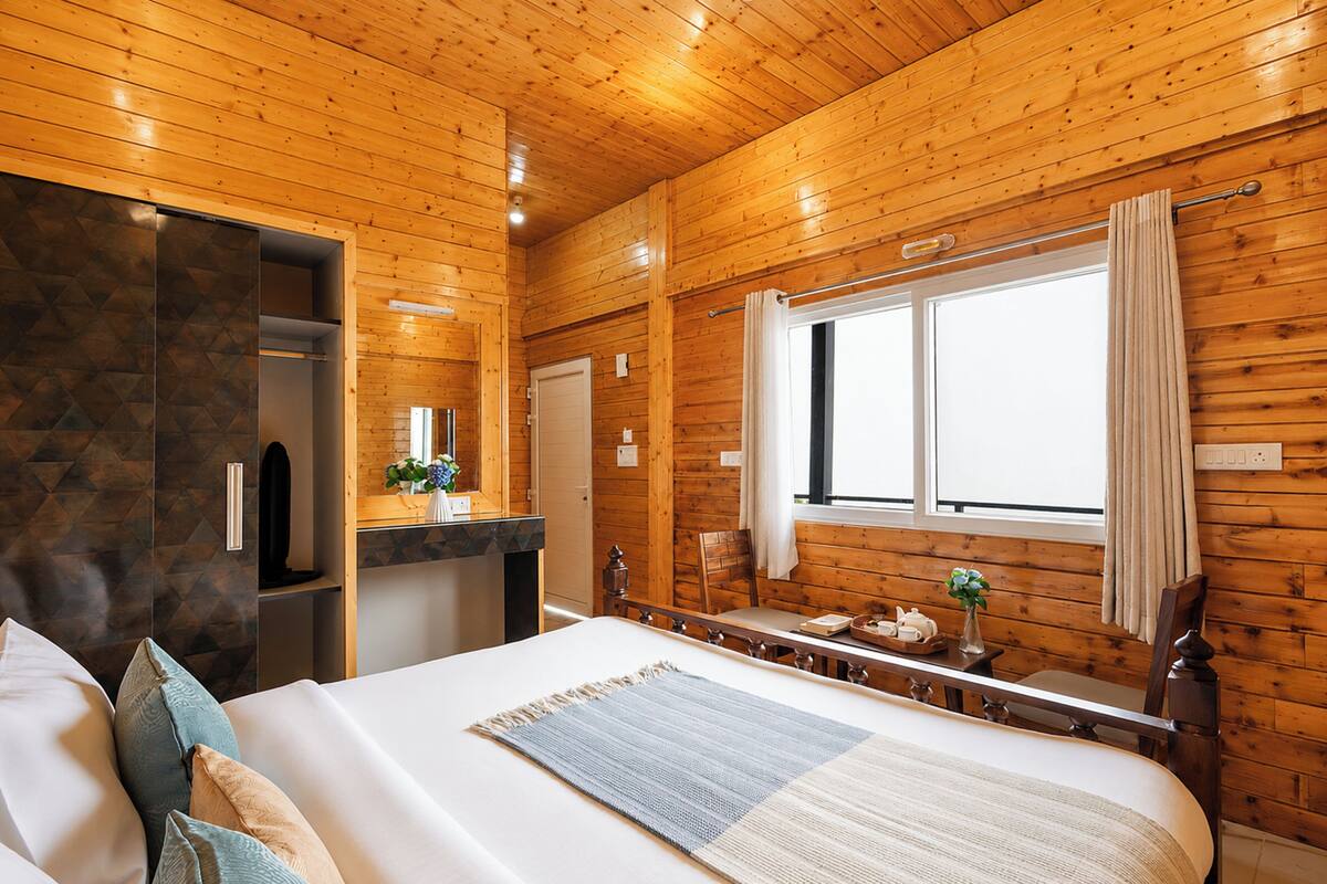 A wooden cabin room with a bed, a vanity, a window with curtains, and a wooden ceiling.