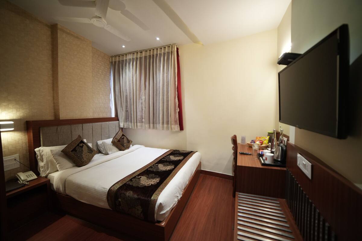 Habitación de hotel con cama, mesita de noche, lámpara, televisión y ventana con cortinas.