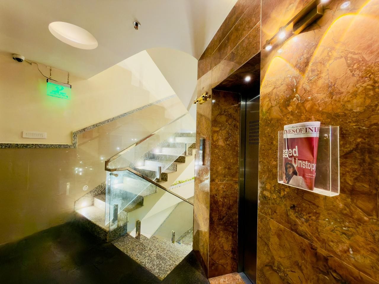 Un interior de hotel moderno con una escalera de cristal, paredes de mármol y un atril con revistas.