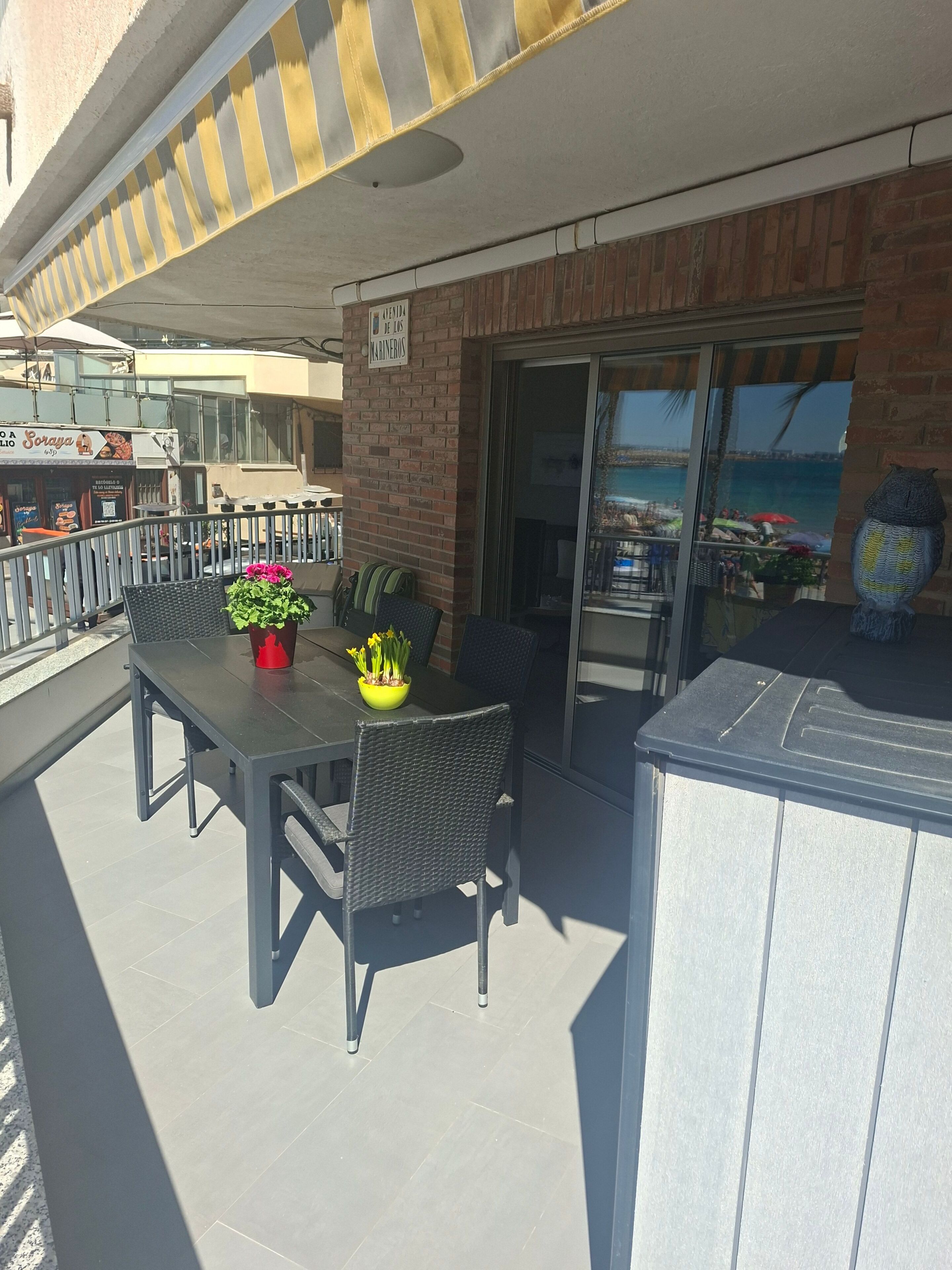 Un patio al aire libre con una mesa y sillas, una planta en maceta y vista a la playa a través de un ventanal.