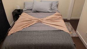 Een bed met een grijze dekbedovertrek en kussens, een beige plaid, een klein nachtkastje met een lamp, en een kleurrijk vloerkleed.