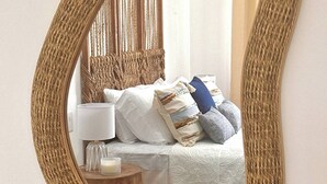 A bedroom with a bed, bedside table, lamp, and a mirror with a woven frame. - CASA RAMONA COSTA RICA (Santa Cruz)