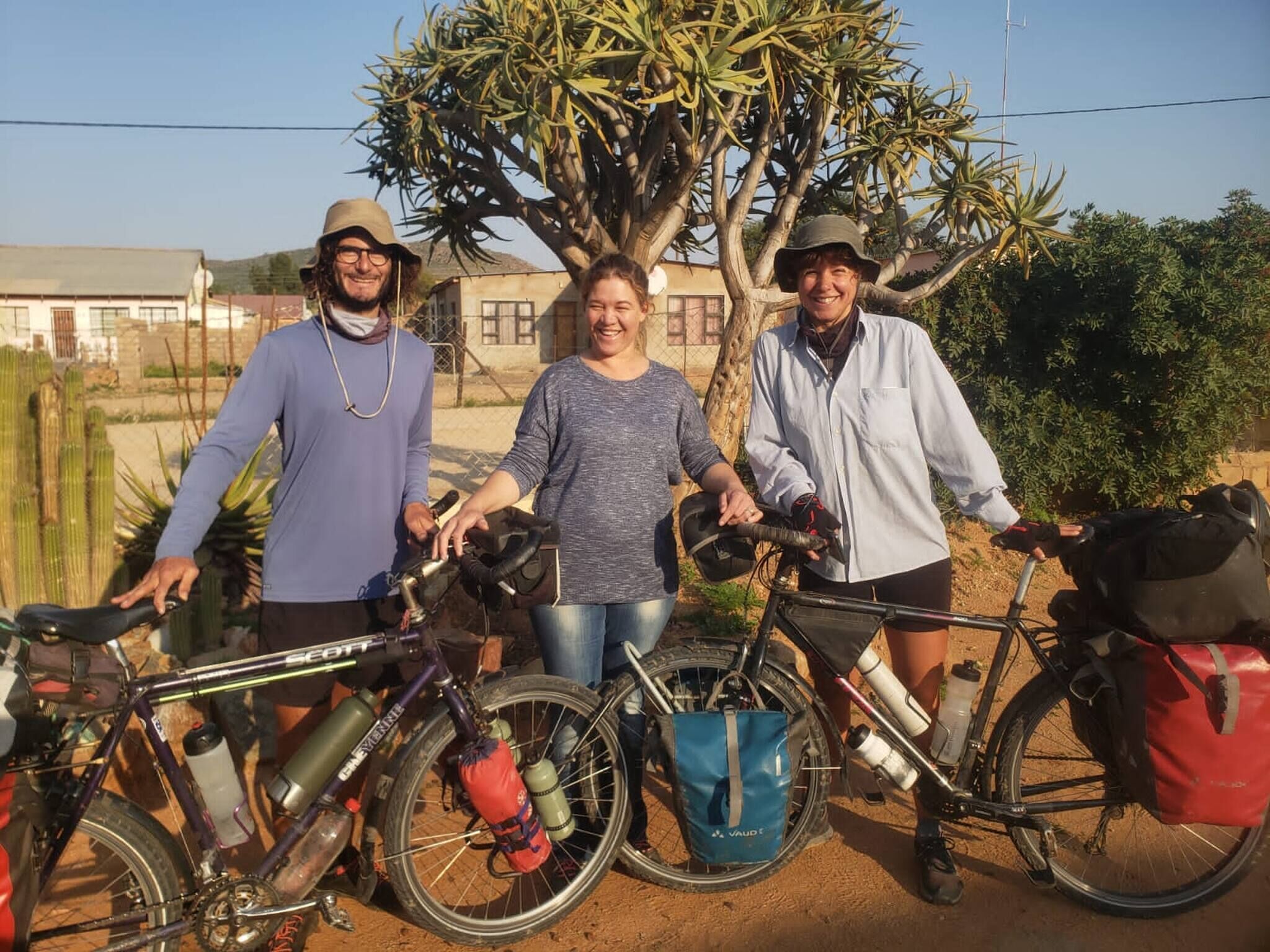 Three people standing with bicycles, a tree, and a building in the background.