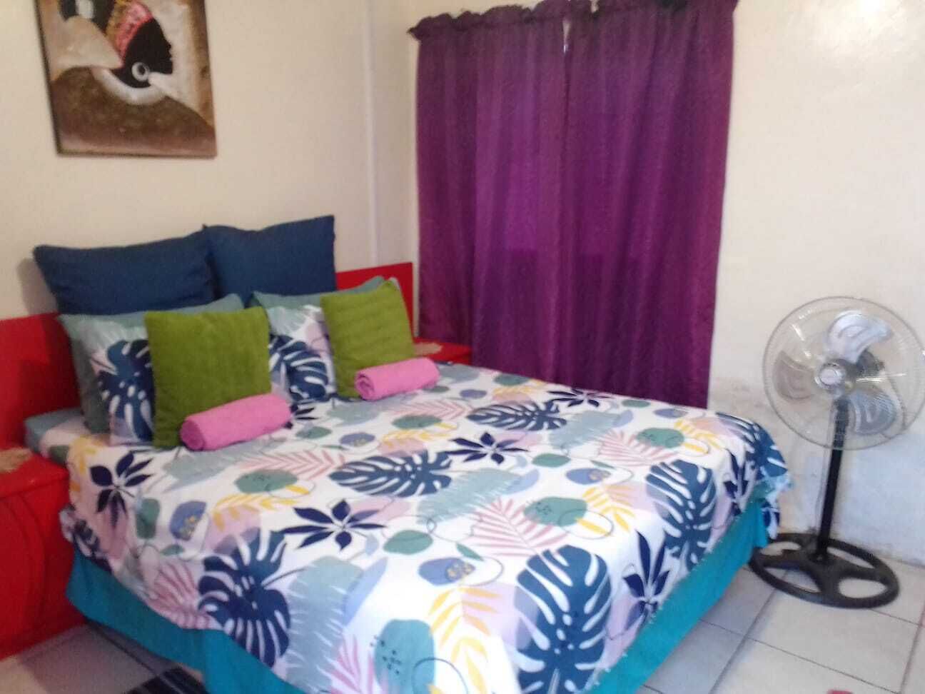 Ein Schlafzimmer mit Bett, Kissen, einem Ventilator und einem Vorhang.