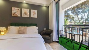Un dormitorio con cama, cabecera verde, mesita de noche con lámpara, balcón con mesa y sillas, y vistas a árboles y edificios.