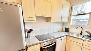 Ett kök med ljusa träskåp, ett kylskåp i rostfritt stål, en inbyggd induktionshäll, en diskmaskin och ett fönster med utsikt över omgivande byggnader.
