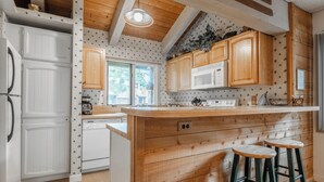 Cucina con mobili in legno, frigorifero bianco, microonde, fornello e due sgabelli da bar.