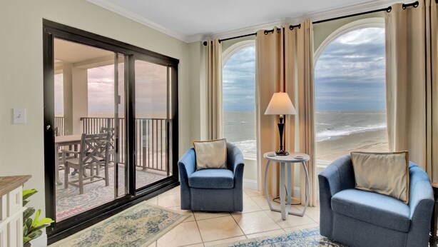 Habitación con vista al océano, dos sillones, una mesita con una lámpara y un amplio ventanal corredera que da a un área exterior con mesa y sillas.