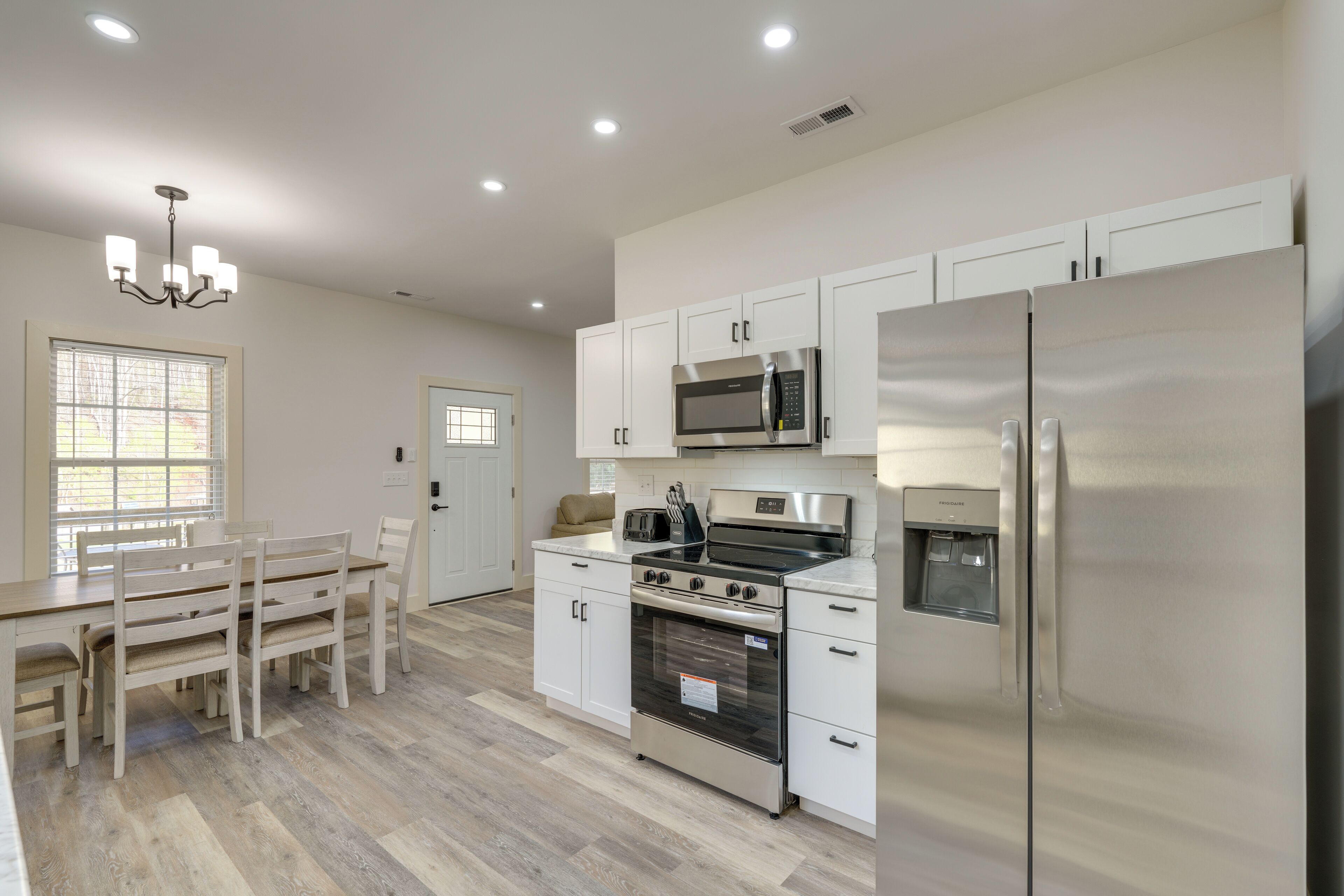 Una cocina moderna con armarios blancos, electrodomésticos de acero inoxidable, una zona de comedor con mesa y sillas de madera, y una araña.