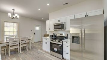 Una cocina moderna con armarios blancos, electrodomésticos de acero inoxidable, una zona de comedor con mesa y sillas de madera, y una araña.