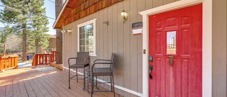A wooden deck with outdoor furniture, a red door, and a window with a view of trees.
