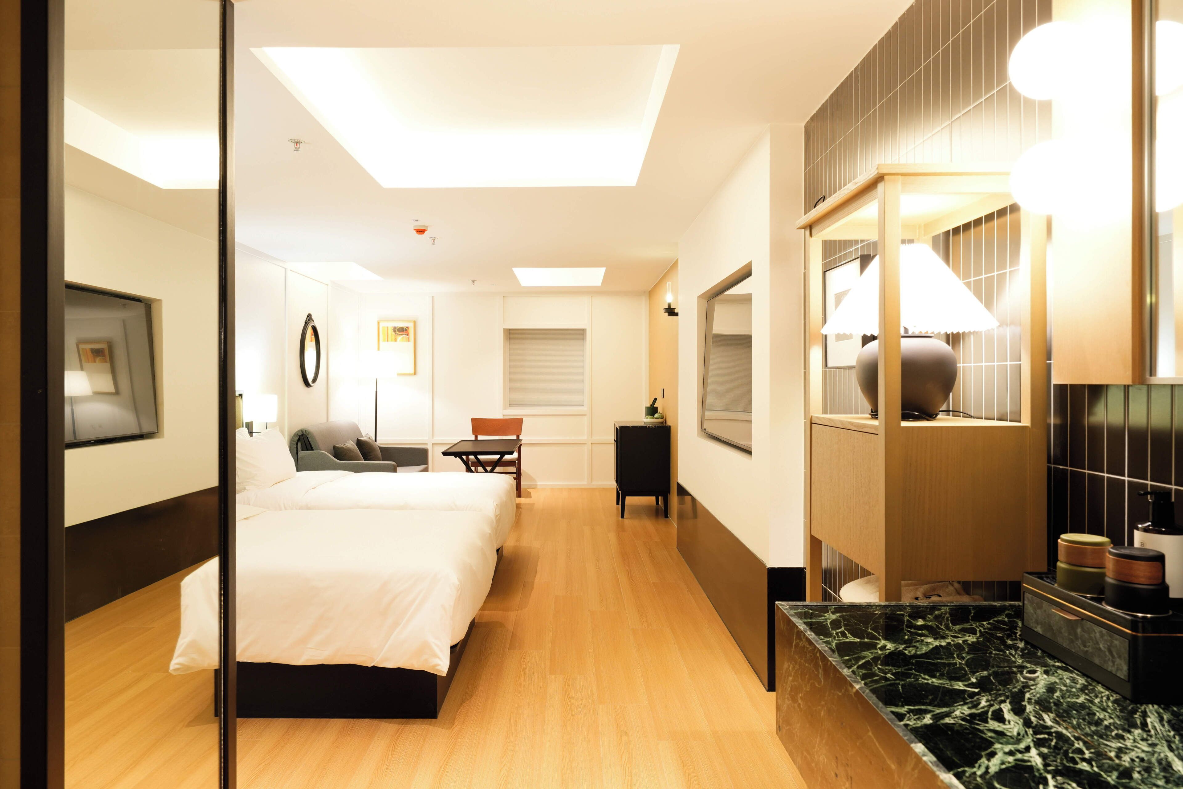 A hotel room with a bed, a desk, a chair, a mirror, and a bathroom with a sink and mirror.