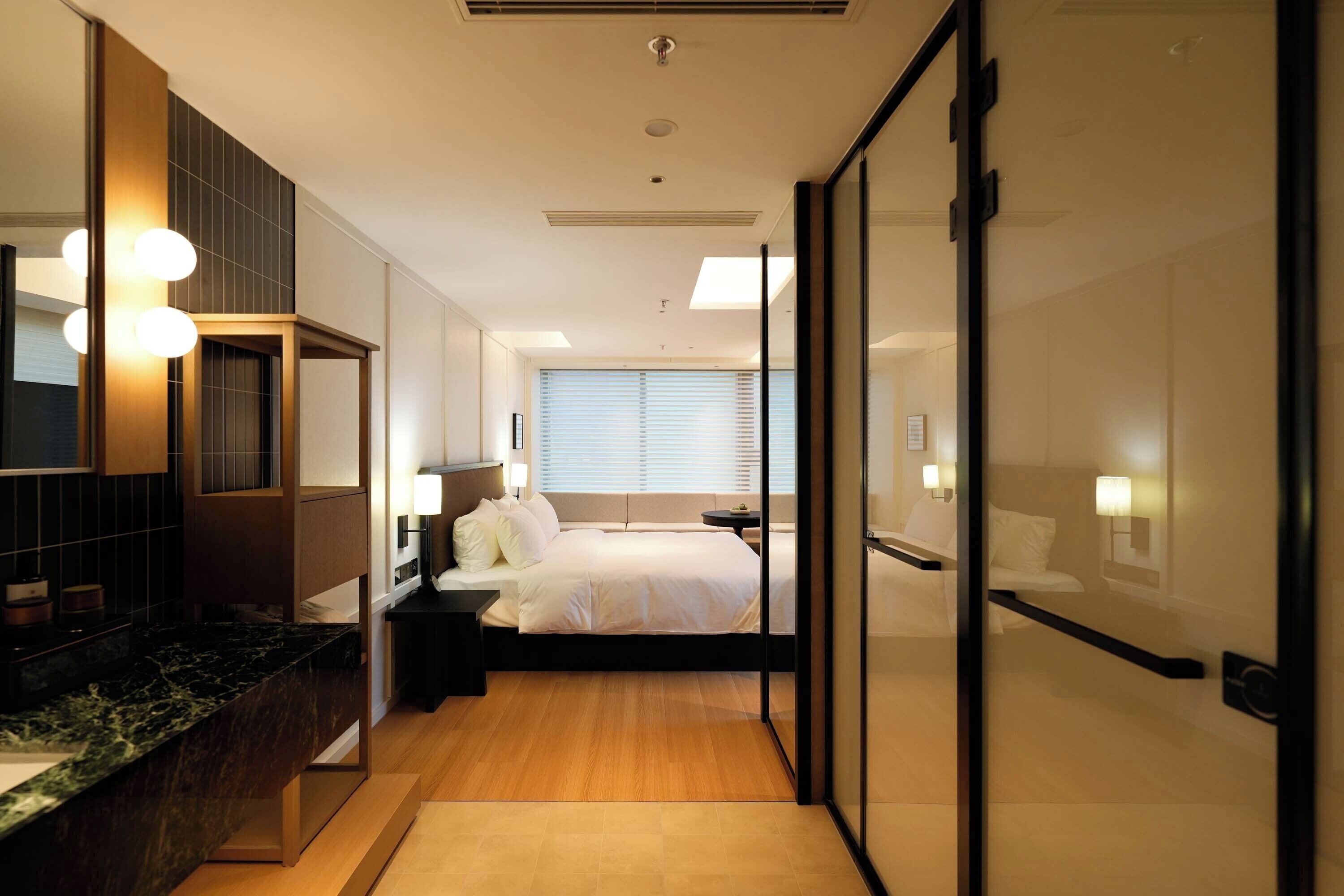 A modern hotel room with a bed, bedside tables, a mirror, and a bathroom with a sink and mirror.