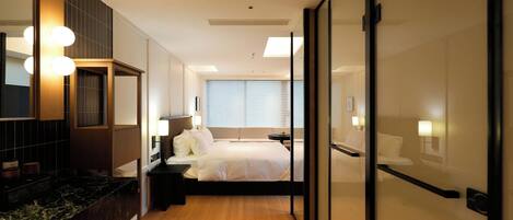 A modern hotel room with a bed, bedside tables, a mirror, and a bathroom with a sink and mirror.