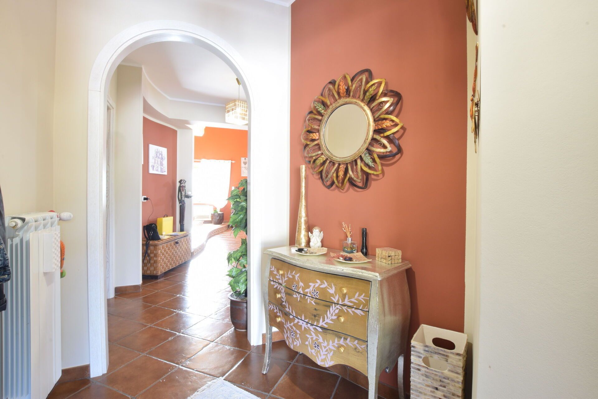 A hallway with a radiator, a decorative mirror, a small table with a vase, and a potted plant.