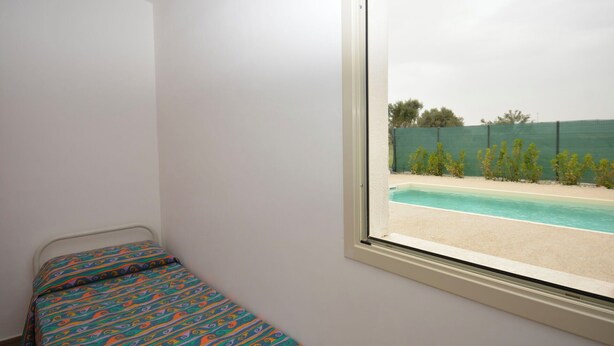 Ein Zimmer mit einem Einzelbett, einem Fenster mit Blick auf einen Swimmingpool und einen grünen Zaun.