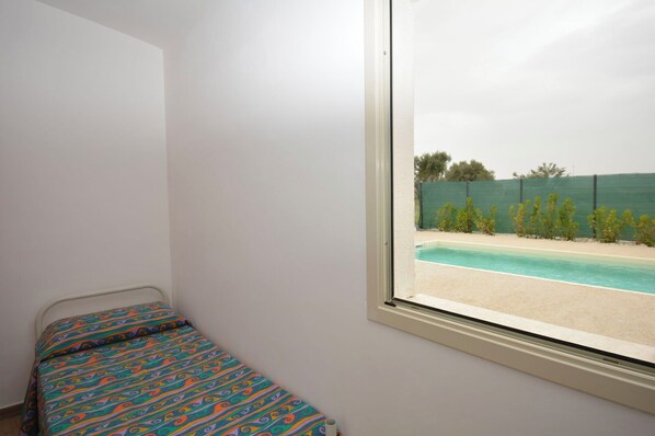 Une chambre avec un lit simple, une fenêtre donnant sur une piscine et une clôture verte.
