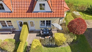Een tweelaags huis met een dak van rode tegels, een terras met een tafel en stoelen, en een verzorgde tuin met verschillende struiken en een rode struik.