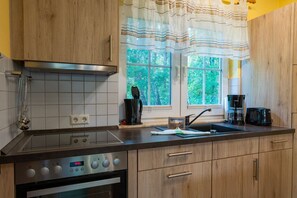 A kitchen with wooden cabinets, a stainless steel stove, a window with curtains, and a coffee maker. - Comfortable vacation home on the dyke with terrace, garden and fireplace in Zingst - Am (Zingst)