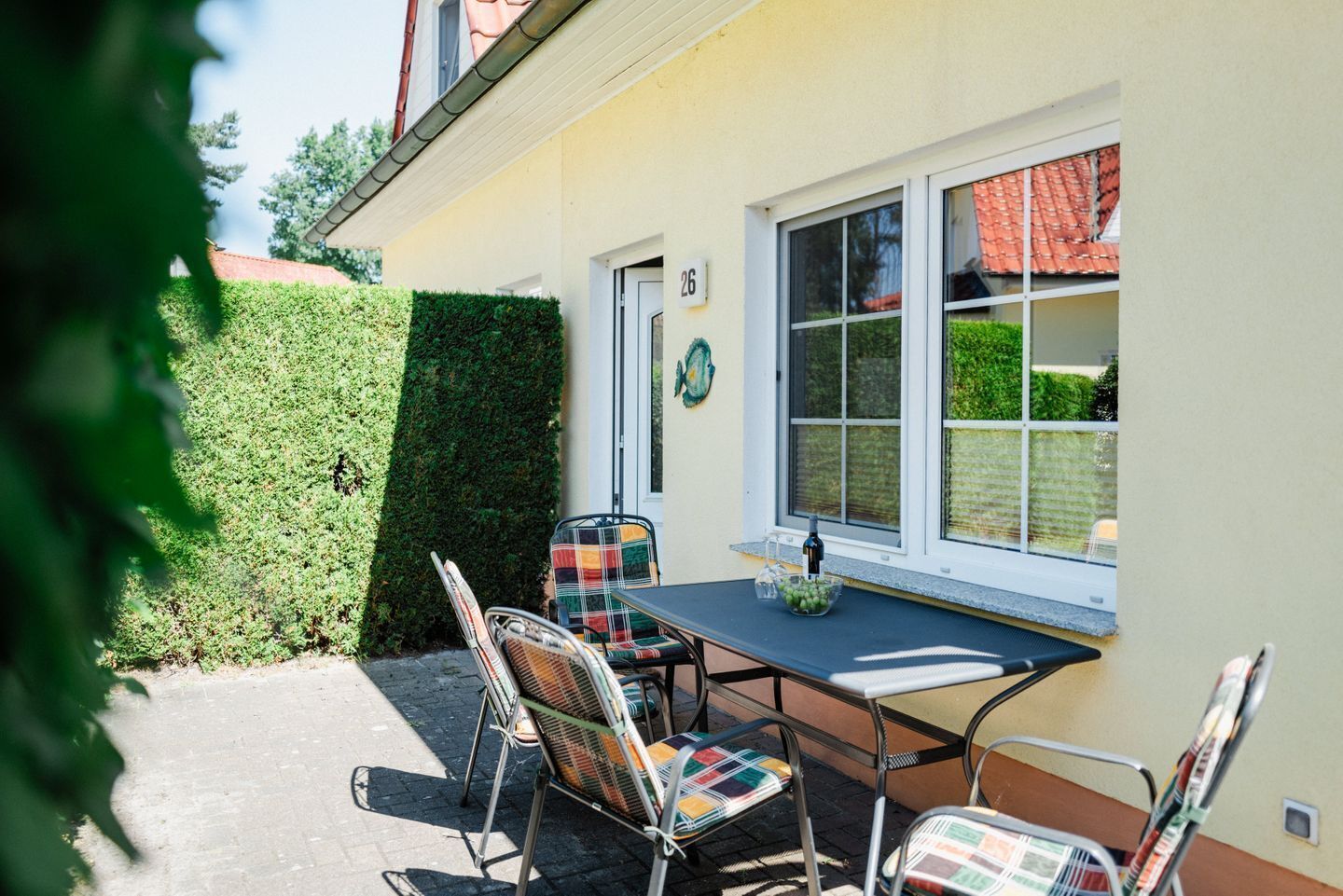 A patio with a table and chairs, a window with a view of a red-tiled roof, and a hedge.