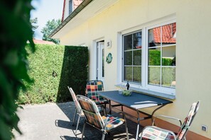 A patio with a table and chairs, a window with a view of a red-tiled roof, and a hedge. - Modern semi-detached house just a few meters from the Baltic Sea beach - Am Deich (Zingst)