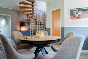Ein Essbereich mit einem runden Tisch, vier Stühlen und einer Fruchtschale. Im Hintergrund ist eine Wendeltreppe zu sehen.