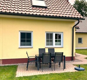 Ein gelbes Gebäude mit einem Ziegeldach, zwei Fenstern und einer Terrasse mit Tisch und Stühlen.