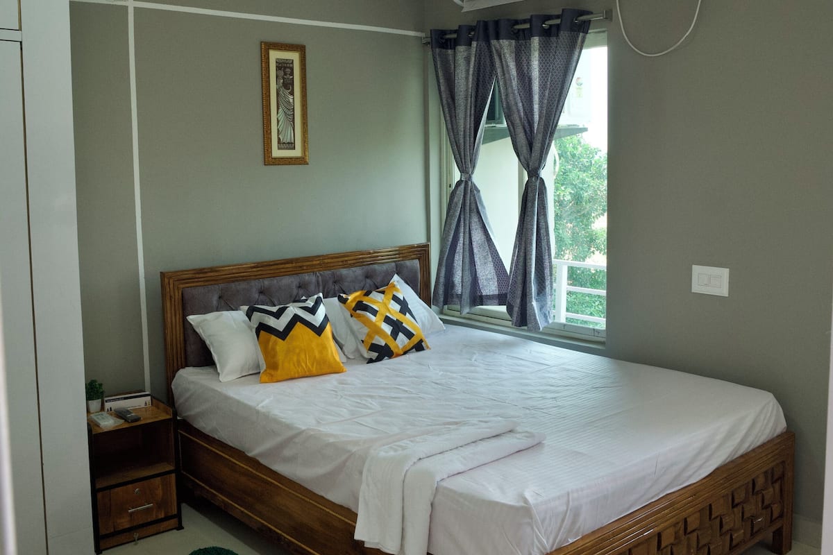 A bedroom with a bed, a ceiling fan, an air conditioning unit, a window with curtains, and a small bedside table.