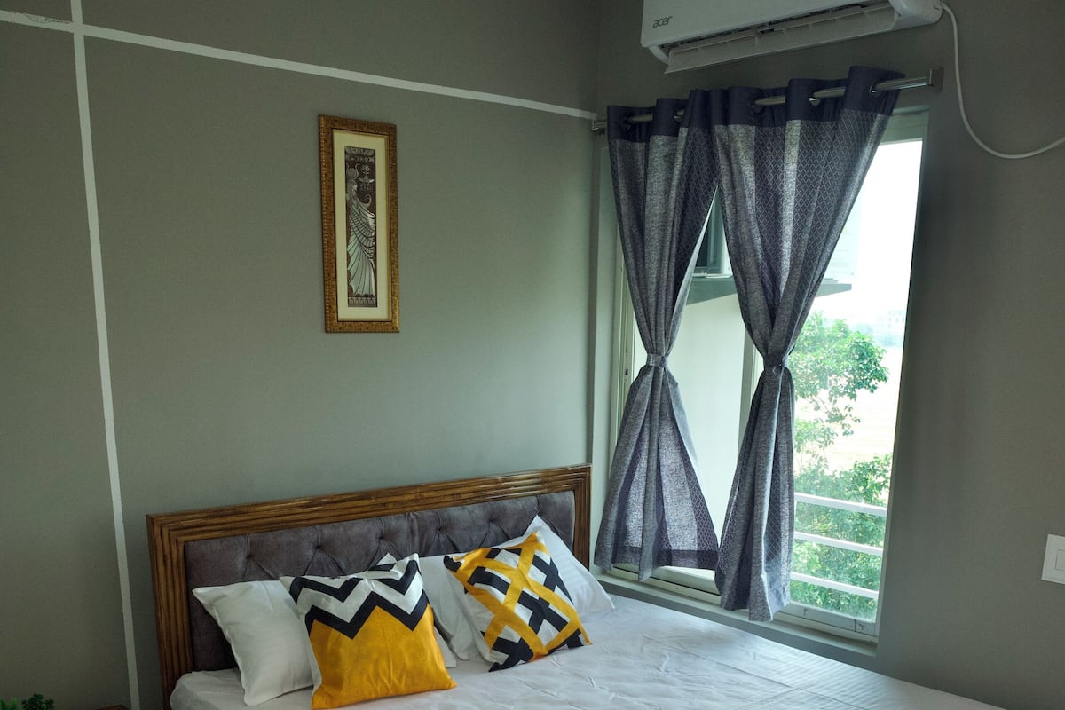 A neatly made bed with a wooden frame, a bedside table with a remote control, and a window with curtains.
