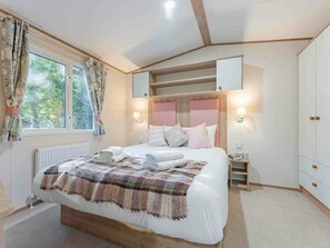 A bedroom with a bed, bedside tables, a window with curtains, a radiator, and a wardrobe. - Mabel - Kelling Heath Holiday Park - 20957 (Holt)