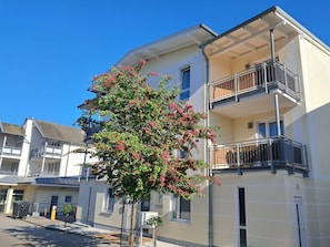 A multi-story building with balconies, a tree with pink flowers, and a clear blue sky. - Central but quiet, balcony, free swimming pool and sauna - Villa Elisenhof Elli 5 (Göhren)