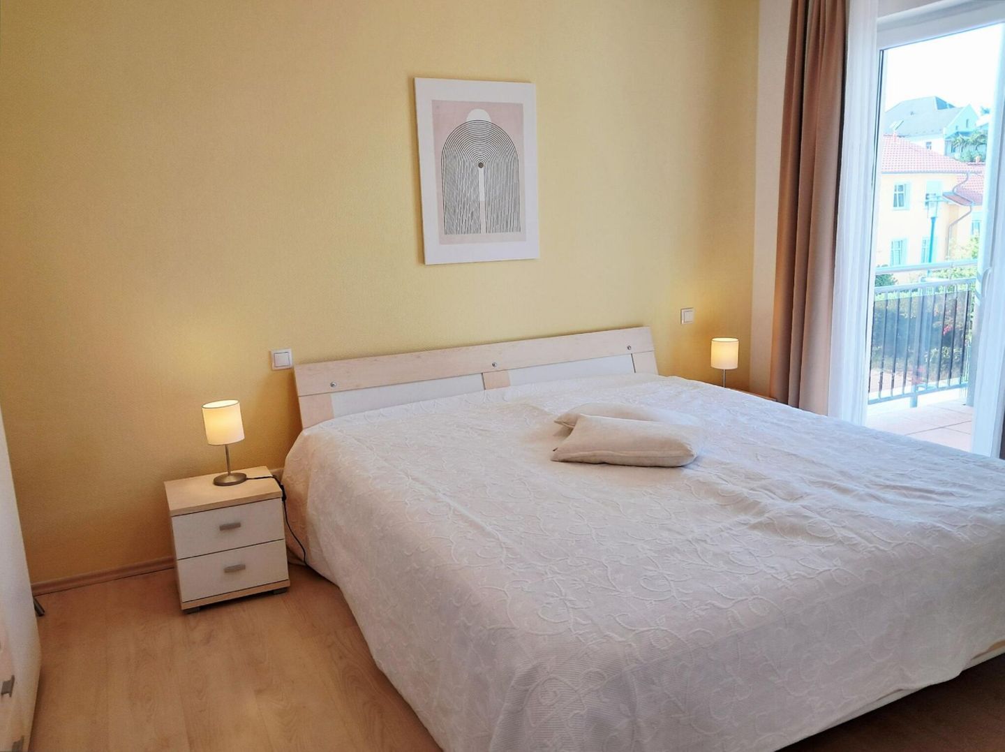 A neatly made bed with a white duvet and pillows, a bedside lamp, a small drawer unit, and a framed artwork on the wall.