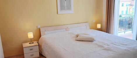 A neatly made bed with a white duvet and pillows, a bedside lamp, a small drawer unit, and a framed artwork on the wall.